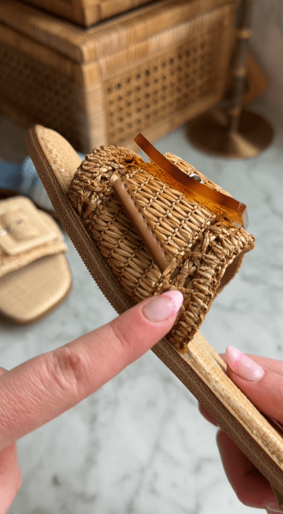 A hand points to the woven detail on a tan, straw-like sandal with a decorative brown buckle, while another similar sandal is visible in the blurred background.