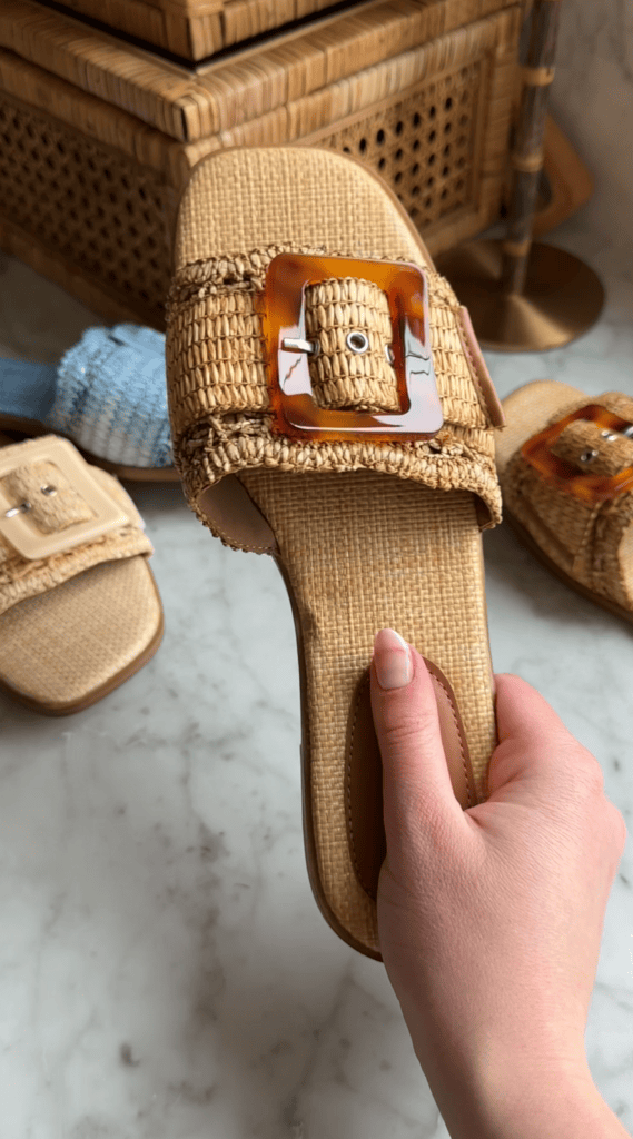 A hand holding a woven sandal with a large brown tortoiseshell buckle, with similar sandals and woven objects blurred in the background on a marble surface.