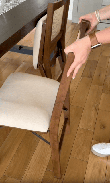 A person wearing a watch and white shoes is unfolding a wooden folding chair with a beige cushion next to a dining table on a wooden floor. Another similar chair is in the background.