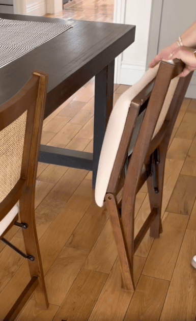 A person is folding a wooden chair with a beige cushion next to a dark wooden dining table on a light wood floor.