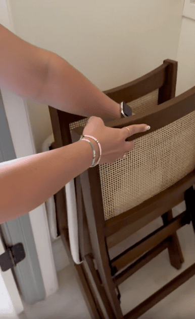 A person with a watch and bracelets on their wrist is holding the backrest of a folded wooden chair with a woven seat, positioned near a wall in a corner of a room.