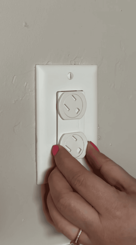 A person with pink nail polish is placing a white safety plug cover into an electrical outlet on a light-colored wall.