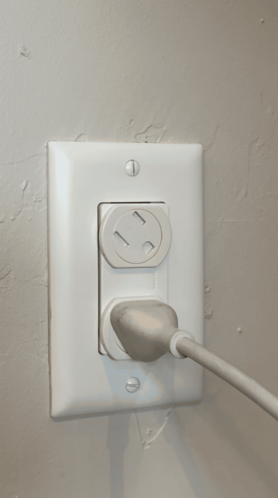A white electrical outlet with a safety cover on the top socket and a white power cord plugged into the bottom socket, mounted on a light gray textured wall.