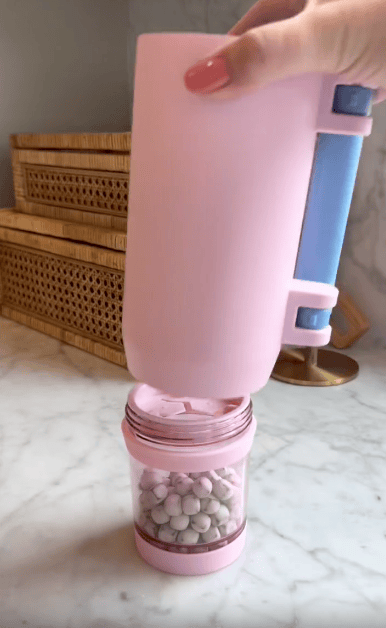 A hand holds a pink Multi Use Snack Tumbler blender with its top section raised, revealing a container filled with small white round objects on a marble countertop, with wicker boxes in the background.