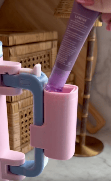 A hand inserts a purple tube into a pink and blue plastic device—possibly a multi use snack tumbler or tube squeezer—with woven baskets and a lamp in the background.