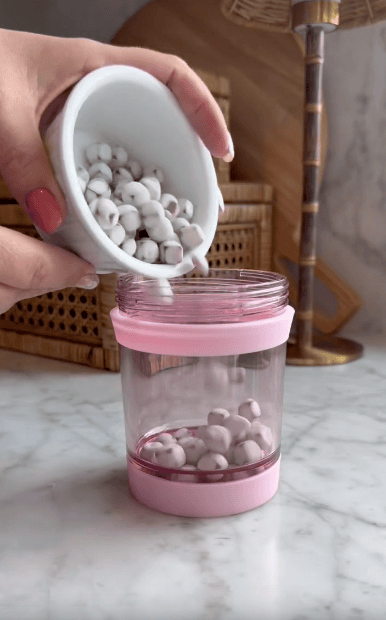 A hand pours white and gray speckled balls from a small bowl into a clear plastic Multi Use Snack Tumbler with a pink lid on a marble countertop. Wicker baskets and a lamp are in the background.