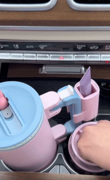 A pastel pink and blue Multi Use Snack Tumbler and organizer set sits in a car’s cup holders. A hand reaches into the organizer, which holds a small tube, while car controls are visible in the background.