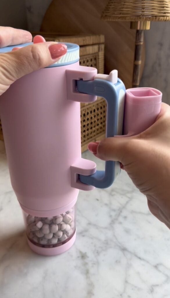 Two hands hold a pastel Snack Tumbler with a handle, twisting the top. The container is filled with small white tablets or candies and sits on a marble countertop.