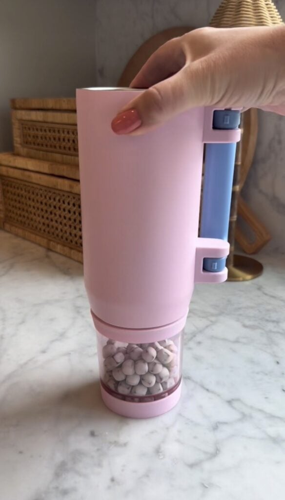 A hand holds a pink and blue Multi Use Snack Tumbler on a marble countertop. The device features small, round white snacks in a transparent compartment. Woven baskets and a lamp are visible in the background.