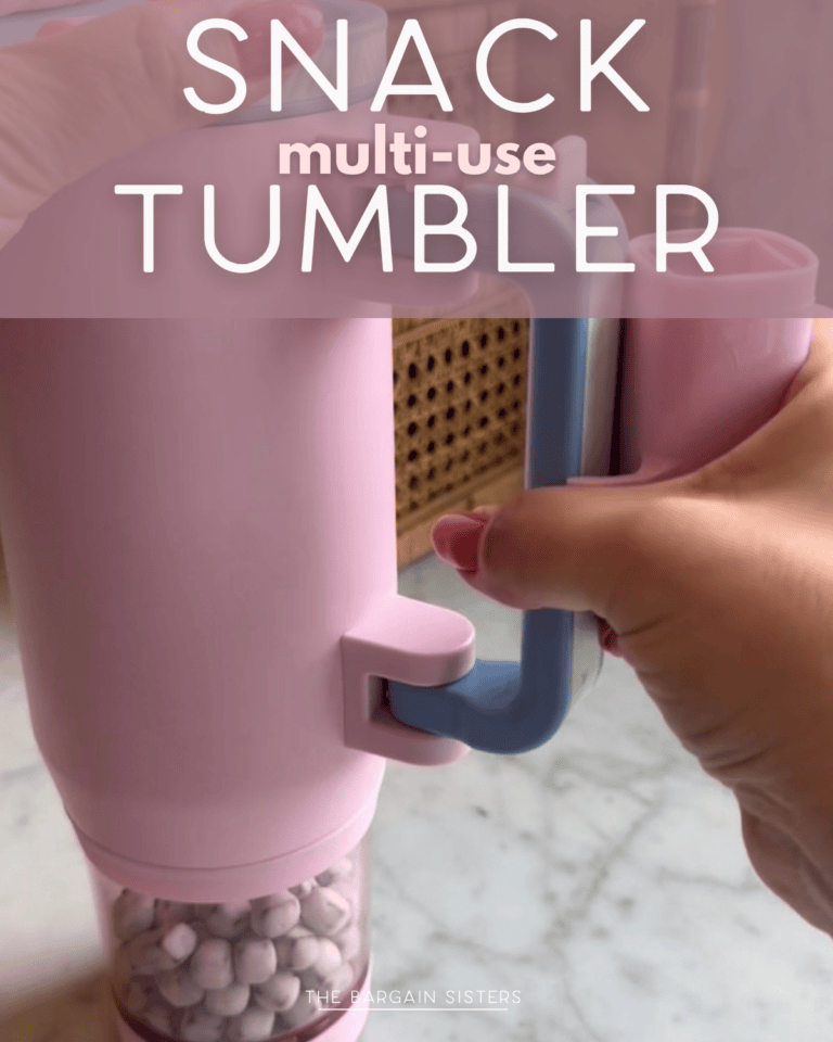 A hand holds a pink Multi Use Snack Tumbler with a blue handle, filled with small snacks. The words "SNACK multi-use TUMBLER" are overlaid on the image. Perfect for snacking on the go, this Snack Tumbler combines style and convenience.