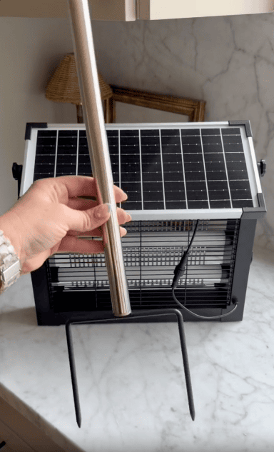 A hand holds a metal rod with a forked end in front of a solar-powered bug zapper resting on a marble countertop.
