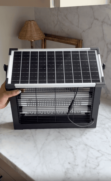 A person holds a black insect zapper with a solar panel attached on top, placed on a marble countertop. A lamp and picture frame are visible in the background.