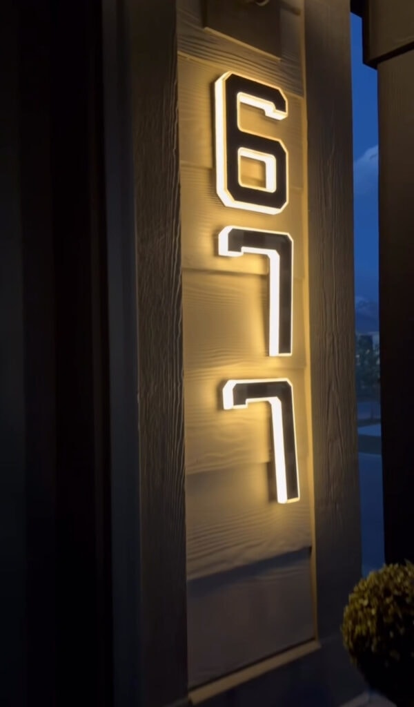 Illuminated house numbers “671” are mounted vertically on a textured wall, glowing warmly against a dark evening background.