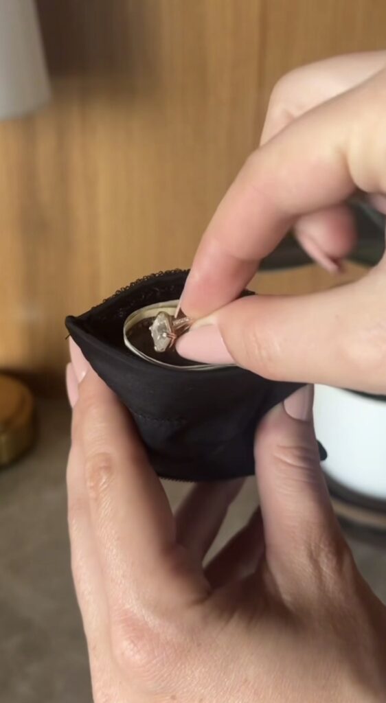 A person holding a black pouch containing a gold ring with a large, clear gemstone, while using their other hand to pick up the ring.