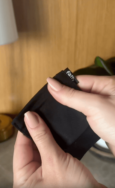 A person with light pink manicured nails holds and folds a small black fabric item with a visible white "fab" label on it, against a blurred wooden background.