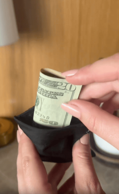 A person with light pink nail polish places a rolled-up $20 bill into a small black pouch. The background shows a blurred wooden surface and some household objects.