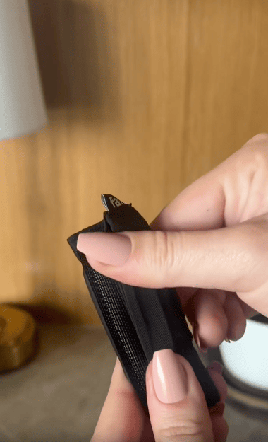 A close-up of a person's hands with manicured nails zipping up a small black fabric pouch, with a wooden surface and blurred objects in the background.