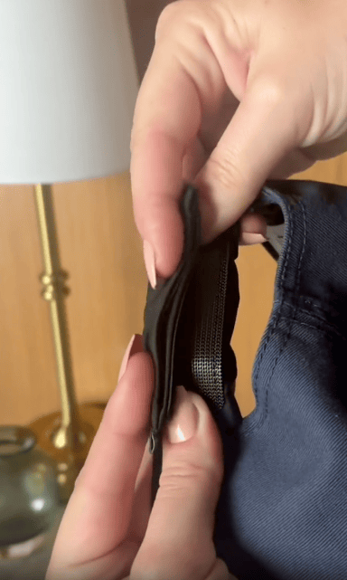 A close-up of hands holding a pair of dark pants, showing a hidden black elastic waistband extension near the zipper. A brass lamp and a glass object are blurred in the background.