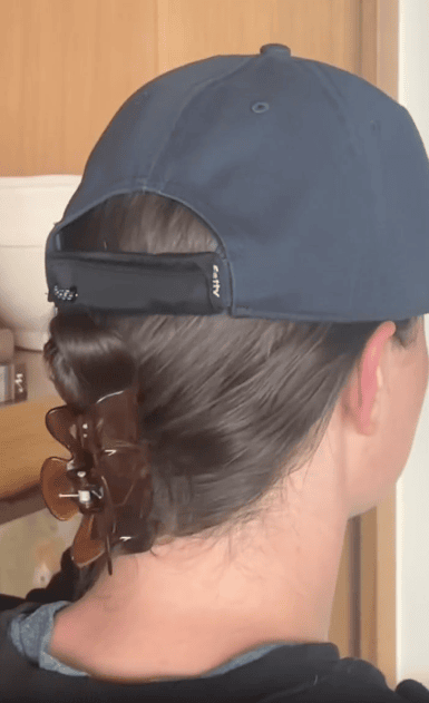 A person with brown hair wears a dark baseball cap backward and uses a large brown hair clip to secure their hair at the back of their neck. The background shows a wooden door and a white bowl on a shelf.