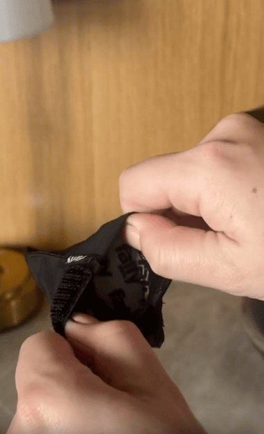 A person’s hands are opening a small black pouch with a Velcro closure, revealing a partially visible label inside. The background includes a wooden surface and a gold-colored object.