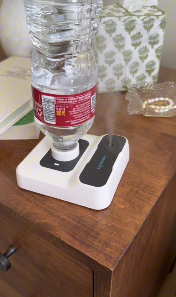 A plastic water bottle is attached upside down to a white tabletop humidifier, placed on a wooden nightstand next to a tissue box, a glass tray with jewelry, and a partially visible book.