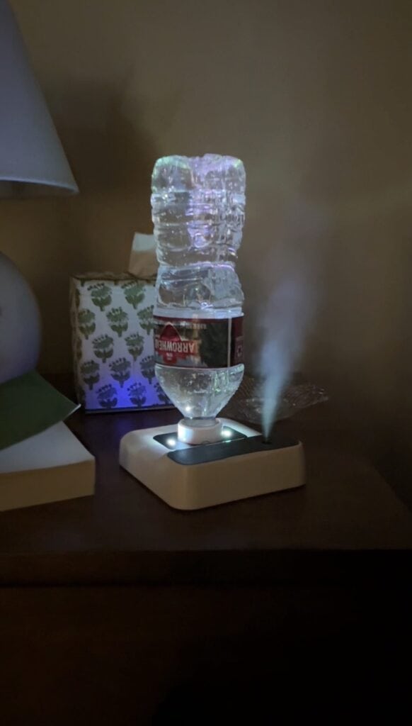 A plastic water bottle is upside down on a small white humidifier next to a tissue box and lamp on a bedside table. Mist is coming out from the humidifier into the air.