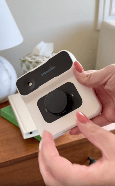 A person holds a white and black electronic device with the brand "Homdis" on it, featuring a large central knob, over a wooden nightstand with books, a lamp, and a tissue box in the background.