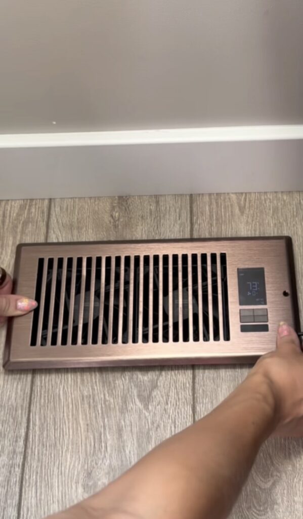Two hands install a bronze-colored vent booster fan with a digital display showing "73°F" on a light wood floor, next to a white baseboard, designed to boost airflow efficiently.