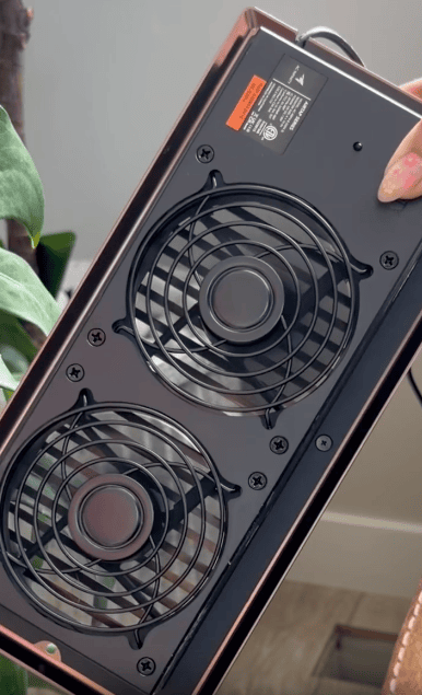 A person holding an electronic device with two black circular cooling fans and protective grilles, designed as a vent booster fan to boost airflow. Visible screws and a label are on the upper corner; green leaves and a white wall are in the background.