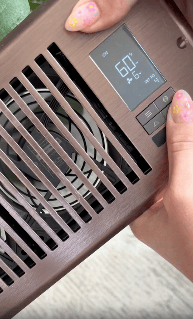 A person with floral nail art adjusts settings on a brown electronic device, displaying 60% humidity, 6°C temperature, and fan level 4 to boost airflow using the built-in vent booster fan.