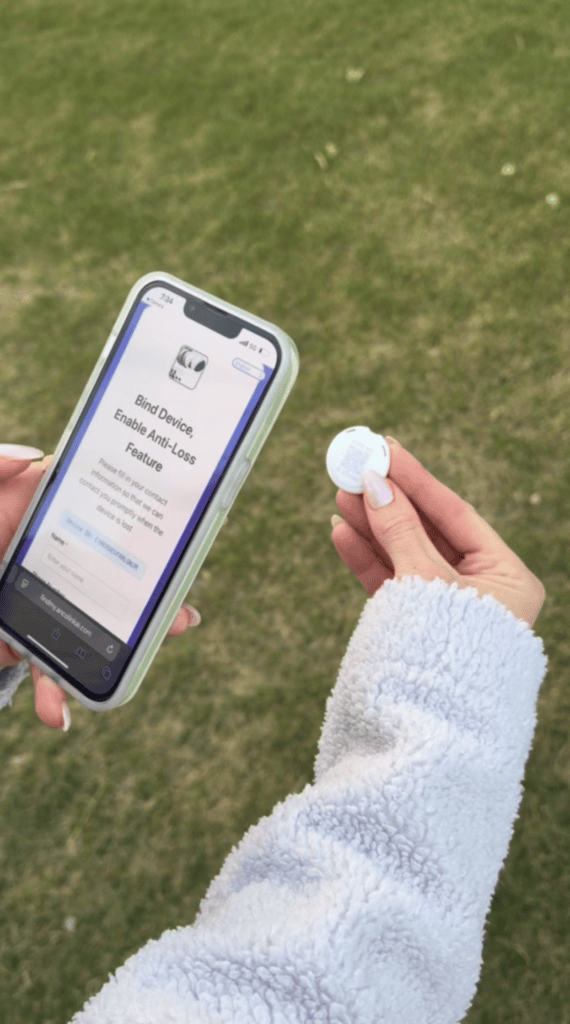 A person wearing a light gray fleece holds a phone displaying a "Bind Device, Enable Anti-Loss Feature" screen and a small round tracking device outdoors on grass.