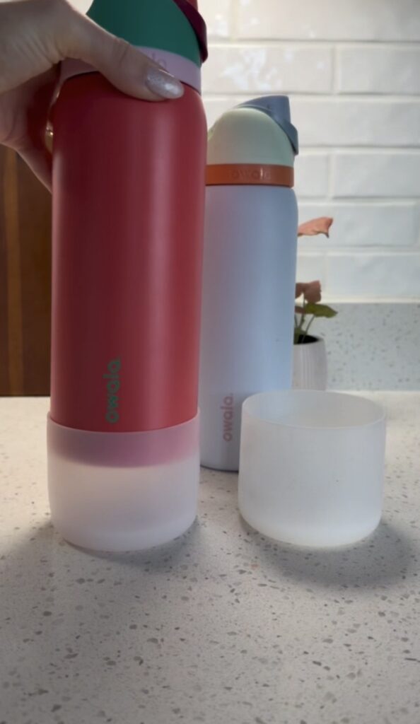 A red and a light blue water bottle with white bases sit on a kitchen counter. The red bottle's base is detached and placed nearby. A hand holds the red bottle, and a small plant is in the background.