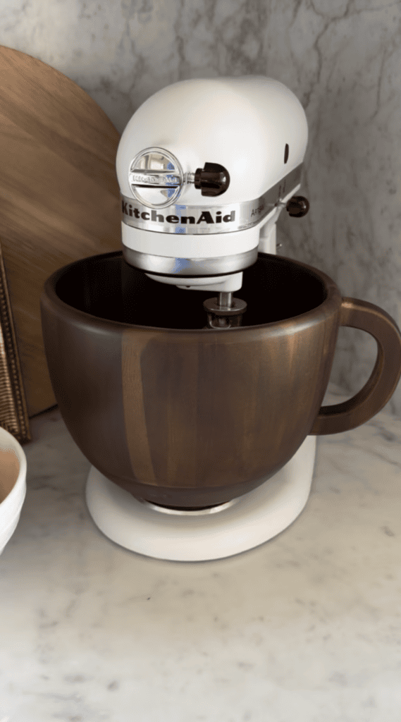 A white KitchenAid stand mixer with a large brown wooden mixing bowl sits on a marble countertop, with a wooden cutting board in the background.