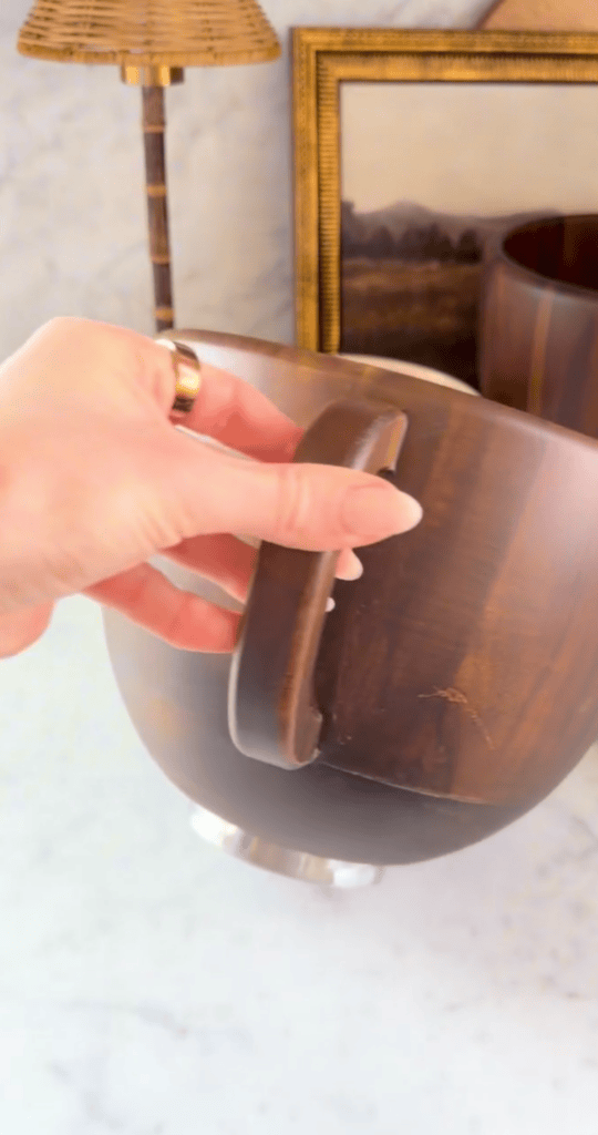A hand holding a wooden bowl with a cracked handle, in front of a lamp, a framed picture, and another wooden container on a marble surface.