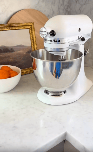 A white KitchenAid stand mixer sits on a marble countertop next to a bowl of oranges and a framed landscape painting.