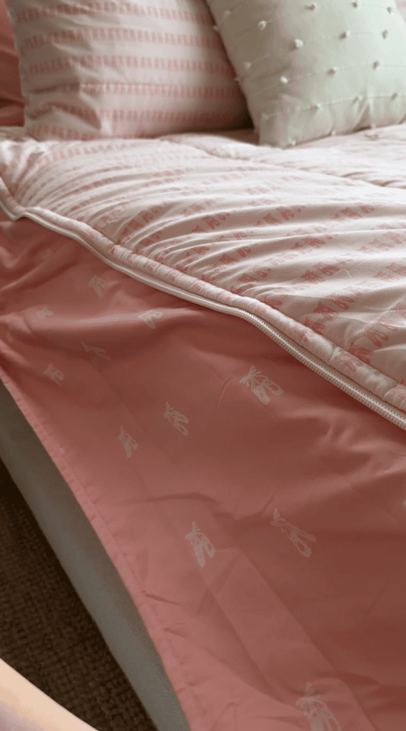 A close-up of a bed with a pink comforter featuring a white pattern, partially unzipped to reveal a matching sheet underneath. The bedding has a soft, cozy appearance and the bed is neatly made.