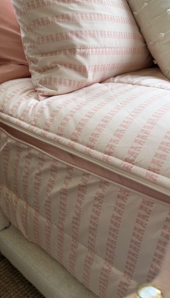 A close-up of a pink and white striped mattress with a visible zipper, topped with matching pillows and bedding. The fabric features repeating small pink designs on a white background.