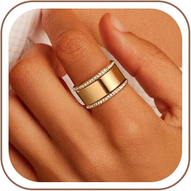 A hand wearing a wide gold ring with two rows of small clear stones bordering the top and bottom edges, set against a soft beige background.