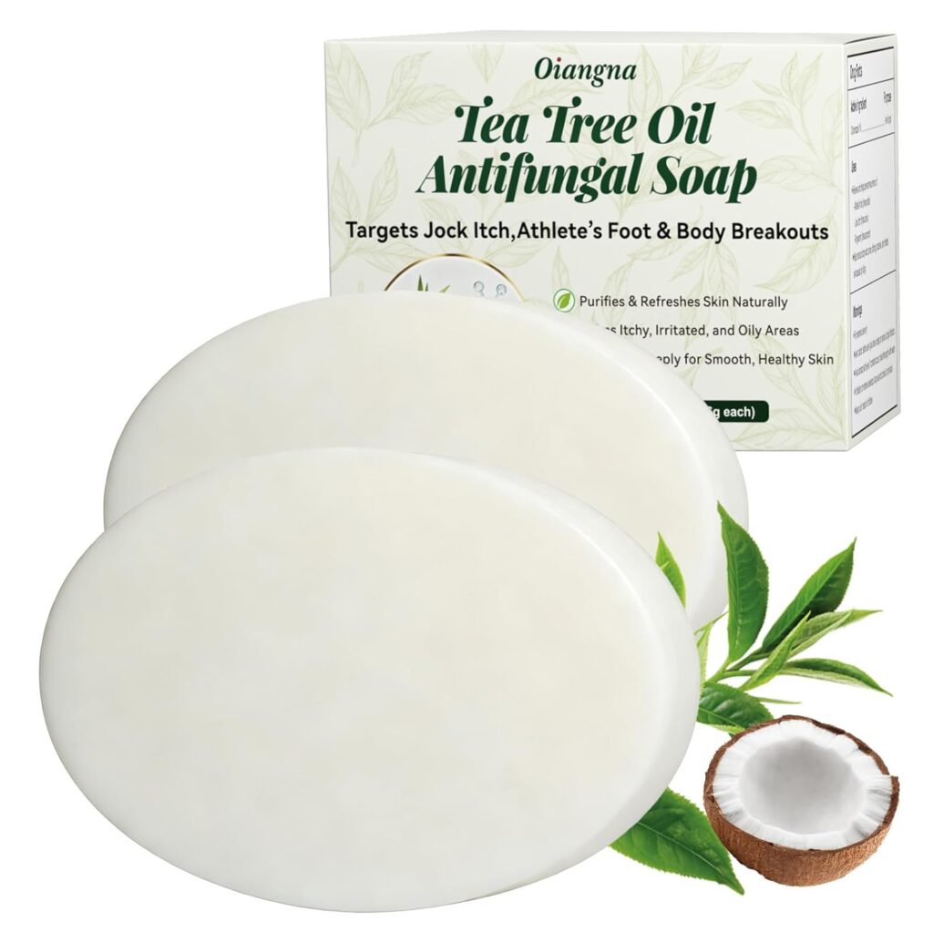 Two oval white soap bars in front of a box labeled "Oiangna Tea Tree Oil Antifungal Soap." Also shown are a coconut half, green leaves, and part of a tea tree branch.