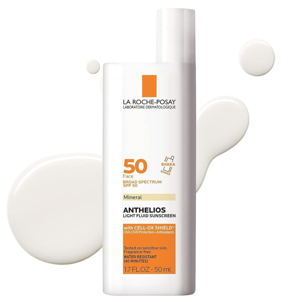 A white bottle of La Roche-Posay Anthelios Mineral Light Fluid Sunscreen SPF 50 is shown against a white background with sunscreen lotion drops around it. The label is orange and white with black and gray text.