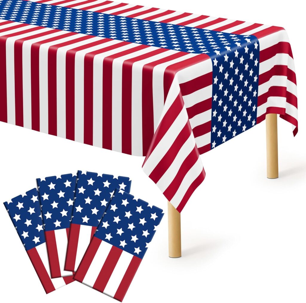 A rectangular table covered with a tablecloth featuring the American flag design, including red and white stripes and a blue field with white stars. Five matching napkins with the same flag pattern are shown in the foreground.