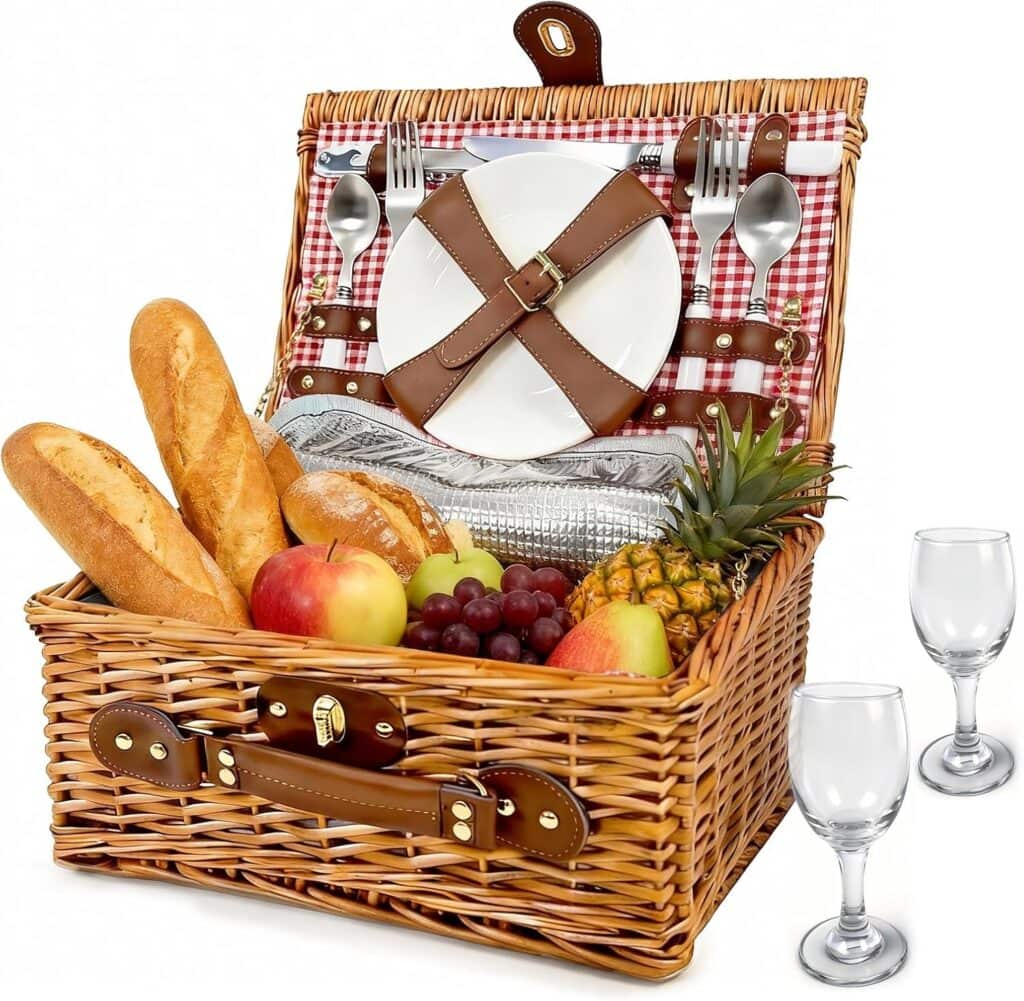 A wicker picnic basket with bread, apples, grapes, pineapple, and cutlery, plates, and glasses arranged inside and beside it, set on a red checkered lining.