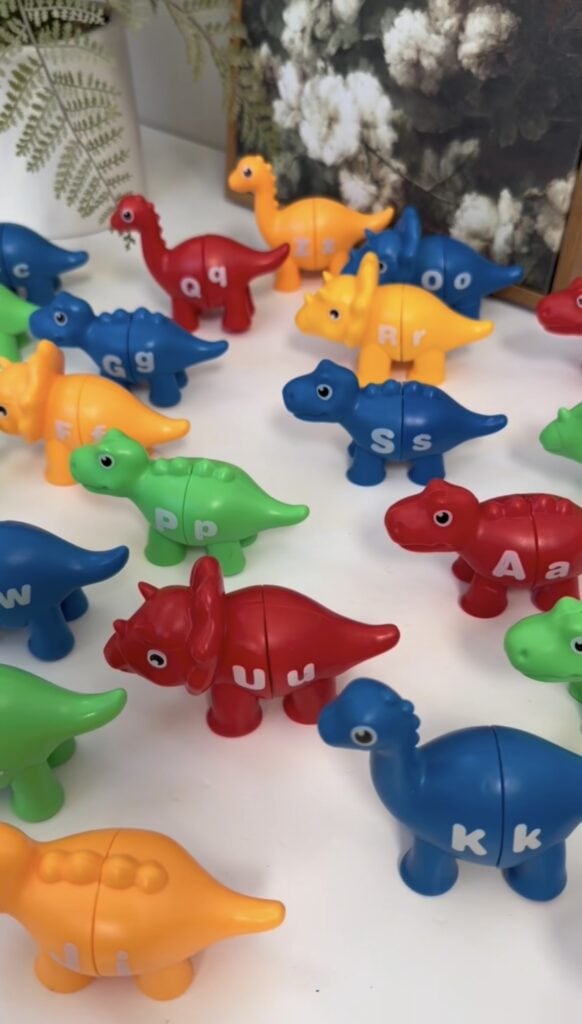 Colorful plastic dinosaur toys are arranged on a white surface. Each dinosaur is divided into two parts and labeled with different letters. Some plants and a blurred picture frame are visible in the background.