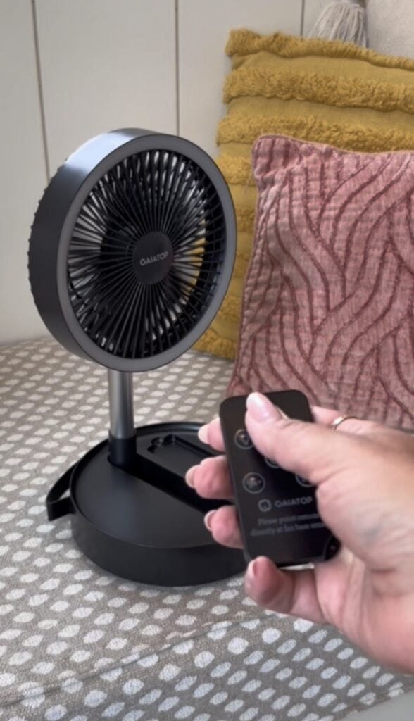 A hand holds a remote control pointed at a small black desk fan on a patterned surface, with pillows in the background.