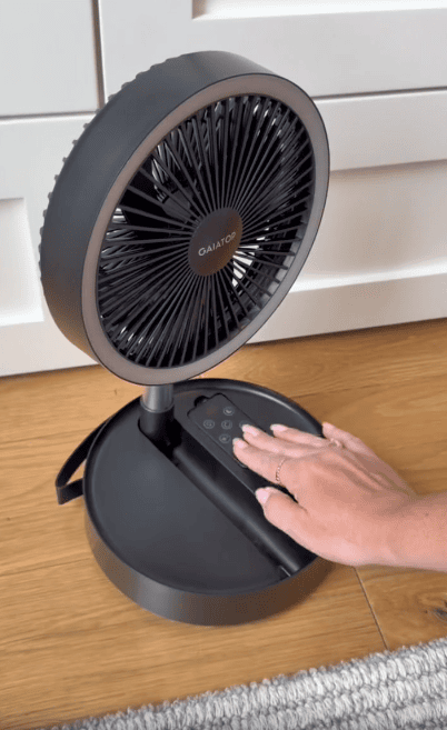A person’s hand presses buttons on a black QAIAOT portable fan placed on a wooden floor in front of white cabinets.