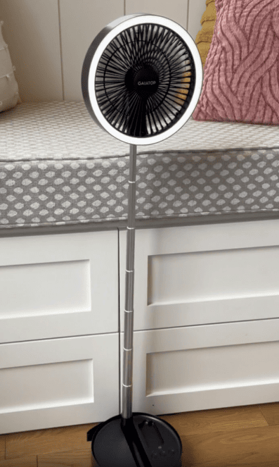 A tall, black and silver standing fan is placed in front of white drawers, next to a cushioned bench with grey polka dot upholstery and a pink textured pillow.