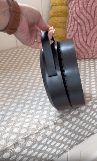 A hand holding a black, round, portable fan with a handle, placed on a beige and white patterned surface. Yellow and pink textured pillows are in the background.