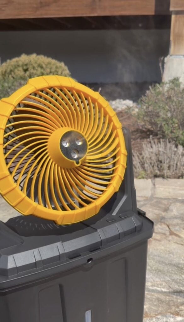 A yellow portable fan is sitting on top of a black plastic bin outdoors, with sunlight shining on it and greenery visible in the background.