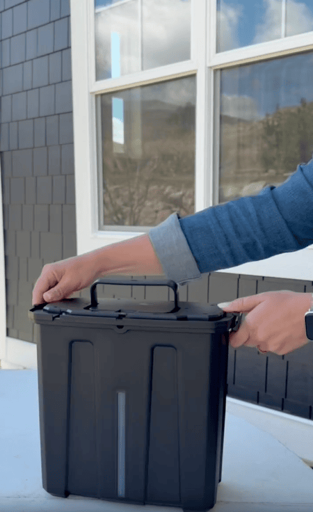 A person wearing a denim jacket and smartwatch unlocks a black, rectangular plastic container with a handle, positioned in front of a window and gray shingled wall.