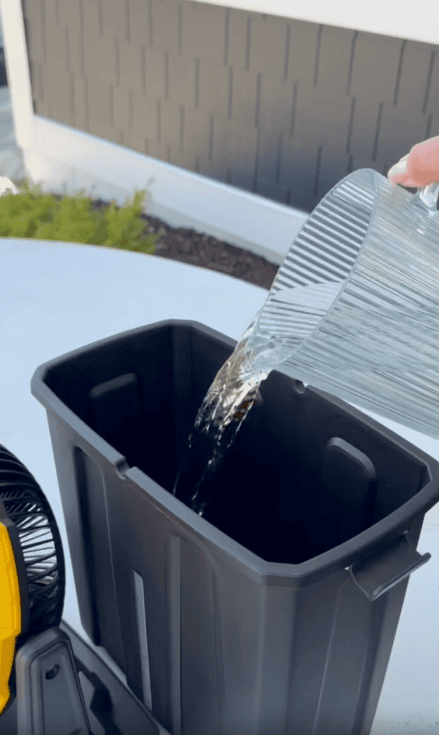 A hand pours water from a glass pitcher into a black rectangular container placed on a white surface outdoors, with part of a fan and a house wall visible in the background.